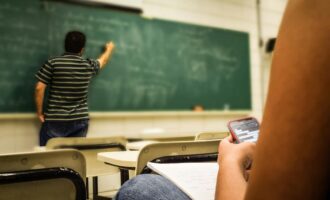 Student texting in a classroom while teacher is writing on the blackboard.