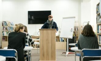 A blind instructor using a cane teaches students in a classroom setting.